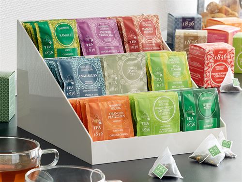 Expositor blanco para tés con diversos tipos de bolsitas de té en un entorno de oficina.
